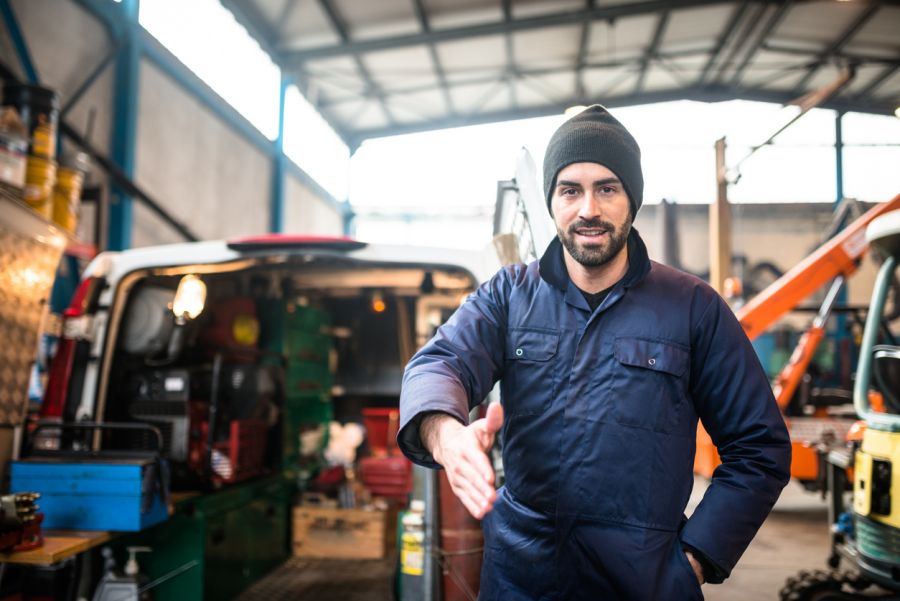 Mobile mechanic in a garage extending a handshake beside a service van and tow truck equipment